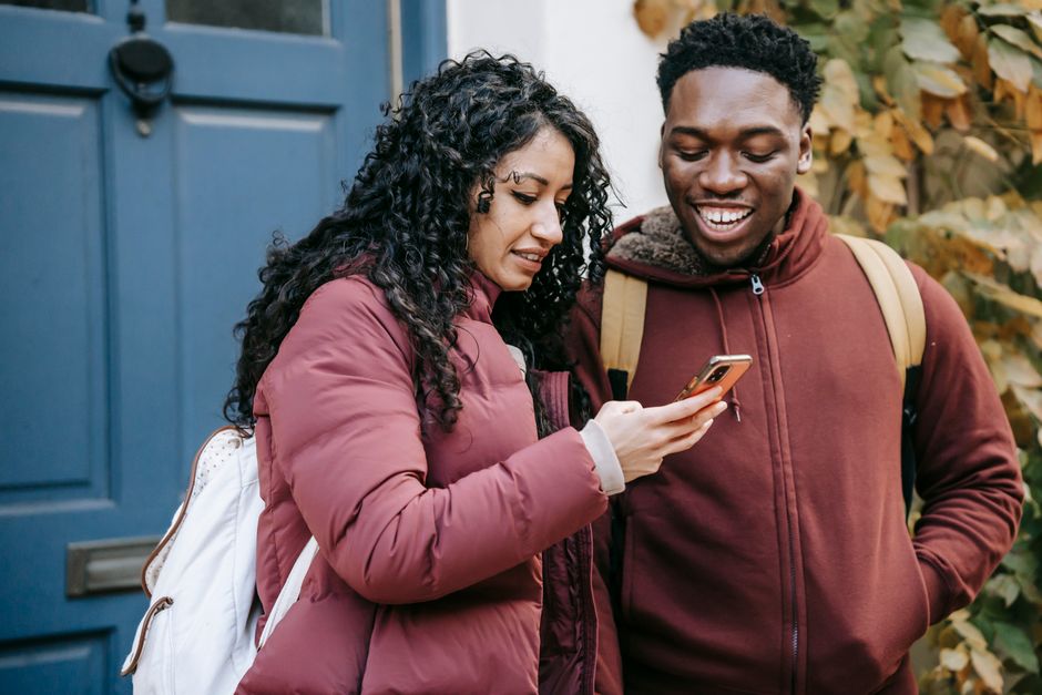 Diverse glad couple in warm outerwear using mobile phone near door of building on street