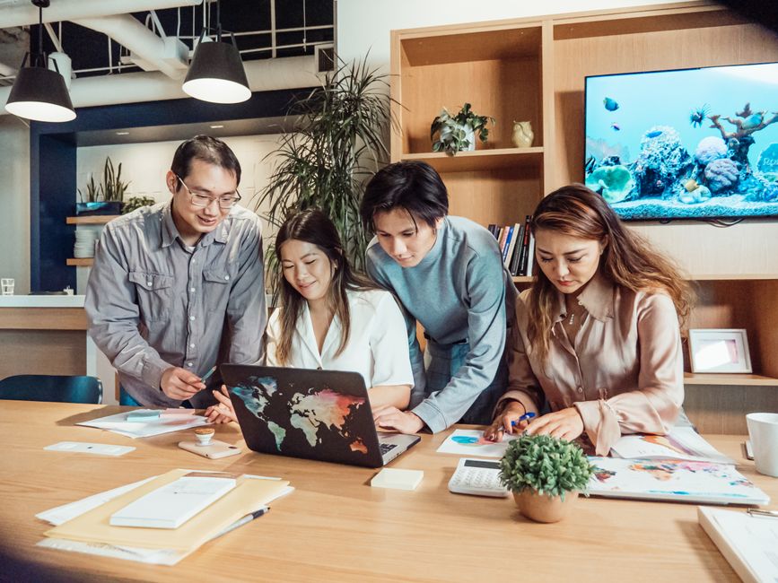 A diverse group of office workers collaborating on a creative project around a laptop in a modern workspace.