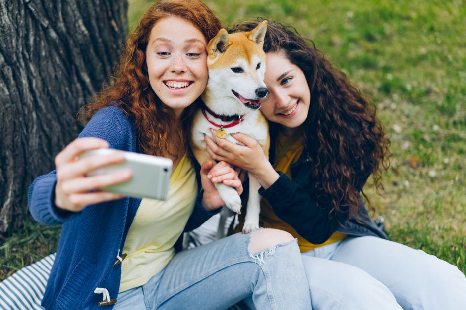 Two women happily taking a selfie with a Shiba Inu dog in a park setting.