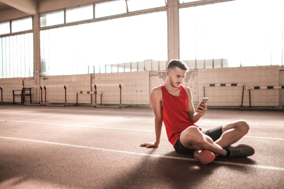 Young male athlete in red sportswear sitting on indoor track using smartphone, embracing modern technology.