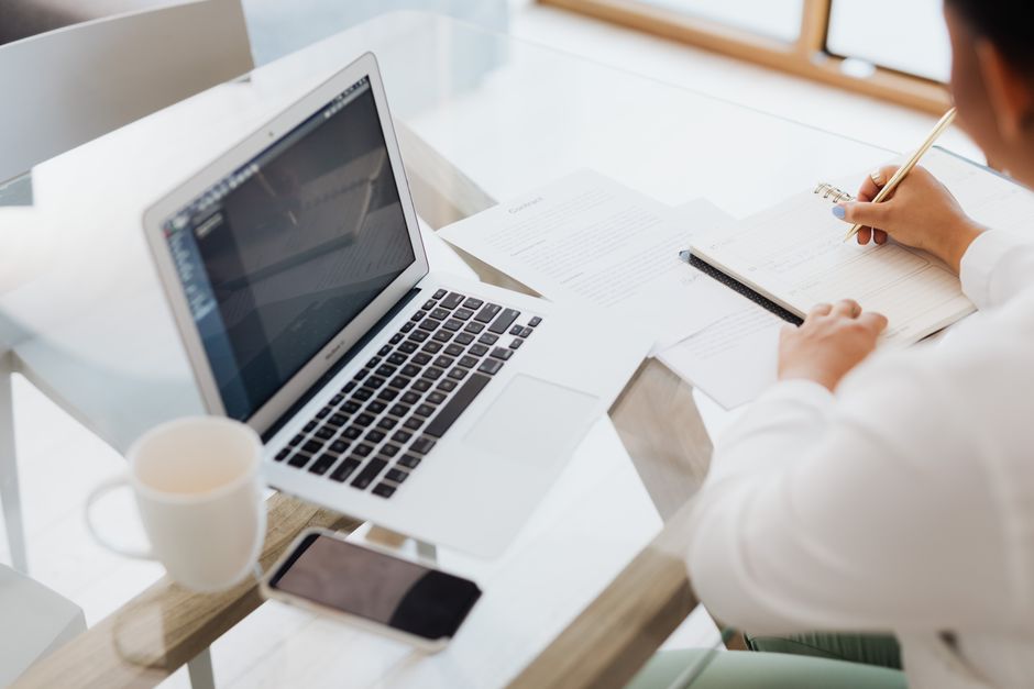 Person working at a glass table with a laptop, notebook, and coffee mug.