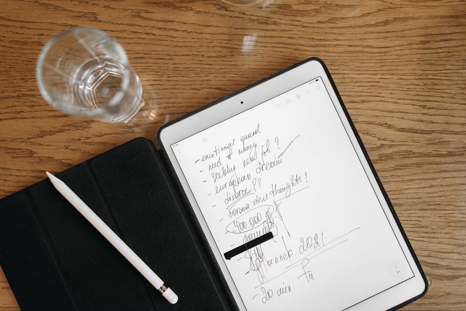 Top-down view of a tablet with handwritten notes, stylus, and glass on a wooden surface.